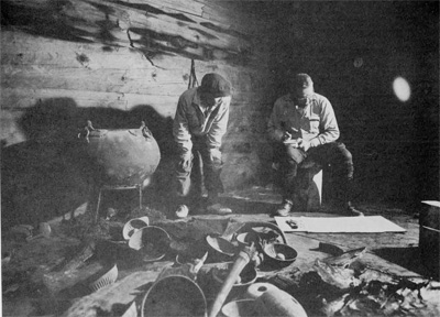 Two men seated in the tomb examining a large paper plan, a variety of bronze objects surrounding them on the ground.