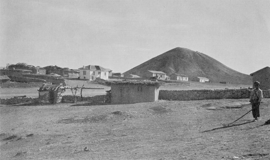 A cluster of small buildings, with a massive mound in the background.