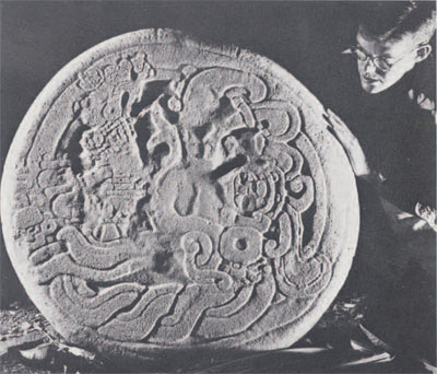 William Coe examines the recently discovered  carving of Altar 12 which depicts an entwined serpent in whose open jaws a priest or deity sits cross-legged holding a glyph and the Maya number 7.