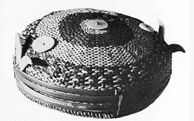 Round, flat basketry hat decorated with four strips of red rattan peel, two pairs of animal teeth, and three shell discs.