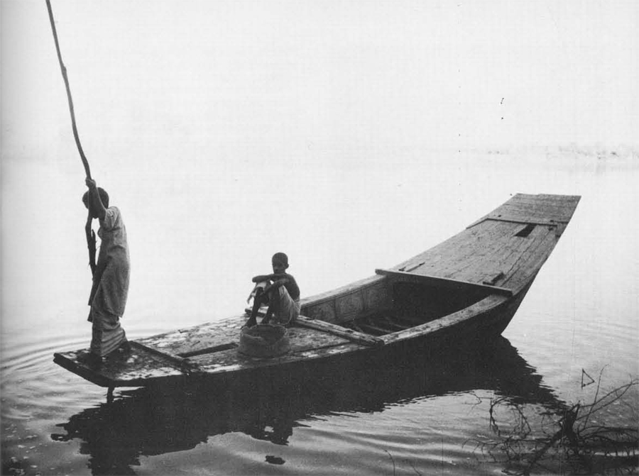 Two people on a wooden boat in the Indus River, one seated, one pushing it along with a long branch.