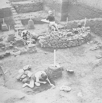 Three people excavating a site, remains of a stone wall can be seen.