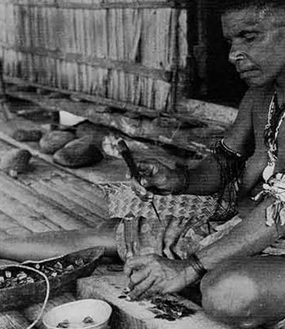 A woman uses a knife handle to break up shell for tambu, a completed string of which lies over the dish in front of her. Her earrings are tortoise shell (but her wristlets are rubber rings from kerosene drums). 
