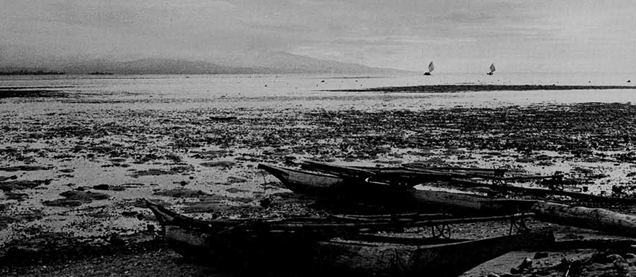 Dugout outrigger canoes beached on an offshore island. Sails of other canoes are visible between the island and the mainland of New Britain. Fresh water, firewood, and all cultivated food except coconuts must be obtained from the mainland.