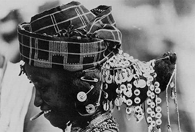 A Fulani women in an ornamented hairdo that culminates in a horn shape protruding from the back of her head, draped in beads, she also wears a headscarf or hat.