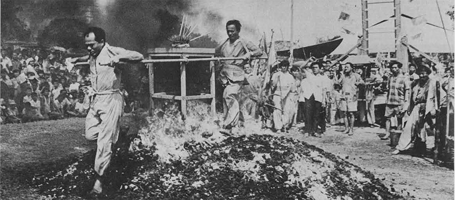 A man walking over hot coals, a line of people waiting behind for their turn to do so, surrounded by onlookers.