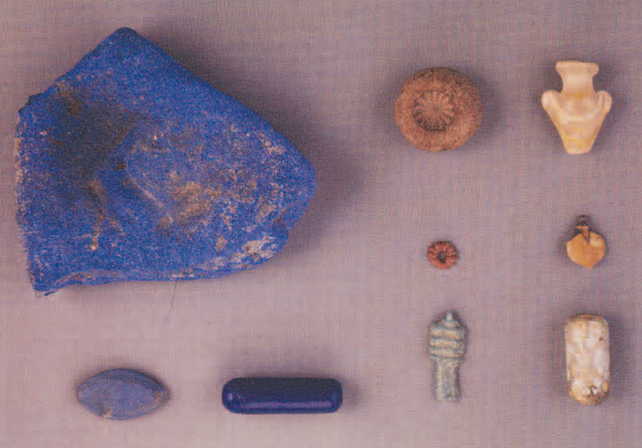 Representative artifacts and manufacturing pieces from the Beth Shan temple and its vicinity. At the left is a fragment of a colorant cake of Egyptian Blue frit (originally a circular flat cake, 8 cms in diameter); below it is a hexagonal ellipsoid bead made of Egyptian Blue frit; the long, dark blue cylindrical bead is of cobalt blue glass. At the top center is a mold for a rosette bead of inlay; the red fluted disc bead below it is of a frit colored with hematite particles; and the dd pendant at the bottom right is of blue-green glazed faience. The heart (ib) pendant at the upper right is decorated with swirls of lead antimonate yellow and calcium antimonate white glass; the mandrake fruit pendant below is of faience overlaid with cobalt-manganese purple and lead antimonate yellow glazes. The cylindrical bead in the lower right corner has cupric blue-green, calcium antimonate white, and lead antimonate yellow crumbs impressed into a silvery-black matrix. 