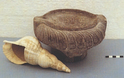 A red limestone lamp decorated with a half foliate band on the shoulder, carved in high relief. It was discovered in Seager's House B along with a cup made by carving out and removing the inside of a triton shell. The two objects were found in a room with a rhyton decorated with palm trees and a clay rhyton shaped like a bull's-head, an exotic assemblage that has suggested to some people that the group represented the contents of household shrine.