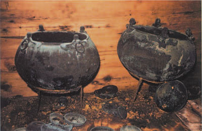 Two large vats or cauldrons which originally contained the mixed fermented beverage of wine, beer and mead, mounted on iron tripod stands. The amphalos drinking bowls in the foreground collapsed off nearby wooden tables.