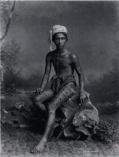 A tattooed burmese villager posing with sword and cigar in the studio of P. Klier, Rangoon, ca. 1898.