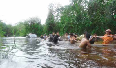 A group of people bulling a truck through a river in two tow lines, holding long ropes.