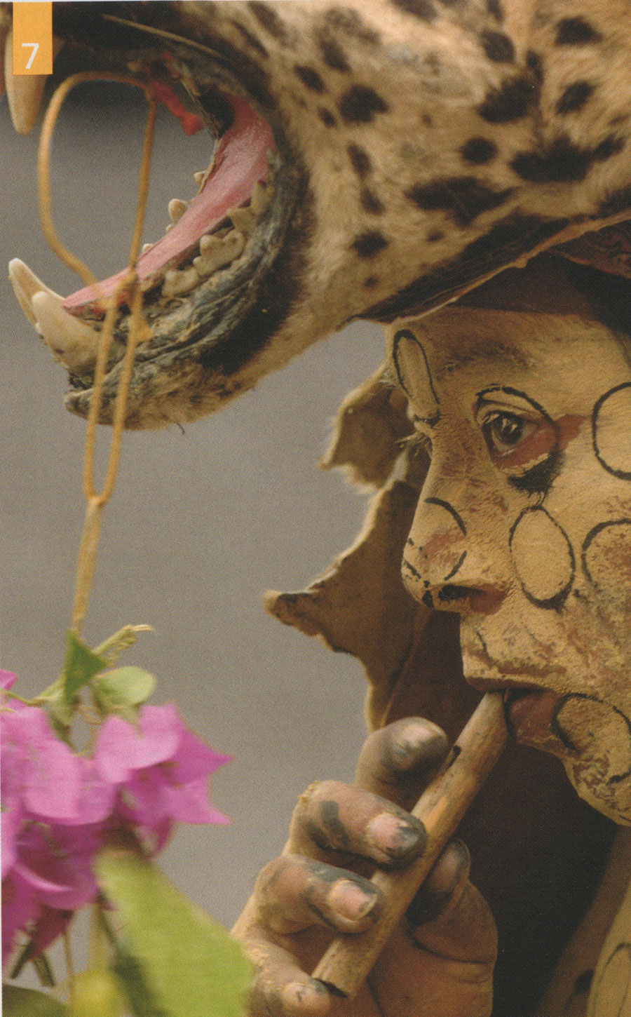 A person in a jaguar headdress and face paint, playing a wood flute.