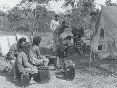 Ainslee Davis sits in foreground with a mixer, just behind a Bororo woman wearing a headset.  in the background, Floyd Crosby and the Bororo woman can see through the lens in the photo to the left) includes George Rawls and a local boy, Tari (UPM image #25661)