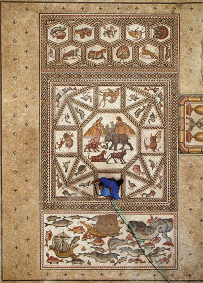 A workman in blue cleans the mosaic. Animals, fish, and birds are depicted along with Roman merchant ships and sea monsters.