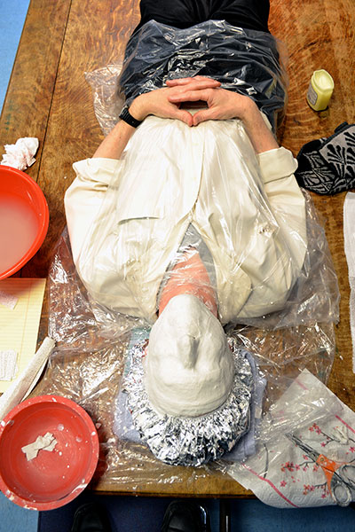 A person laying on a table, plastic wrap covering their head and torso while a plaster mold of the persons face.