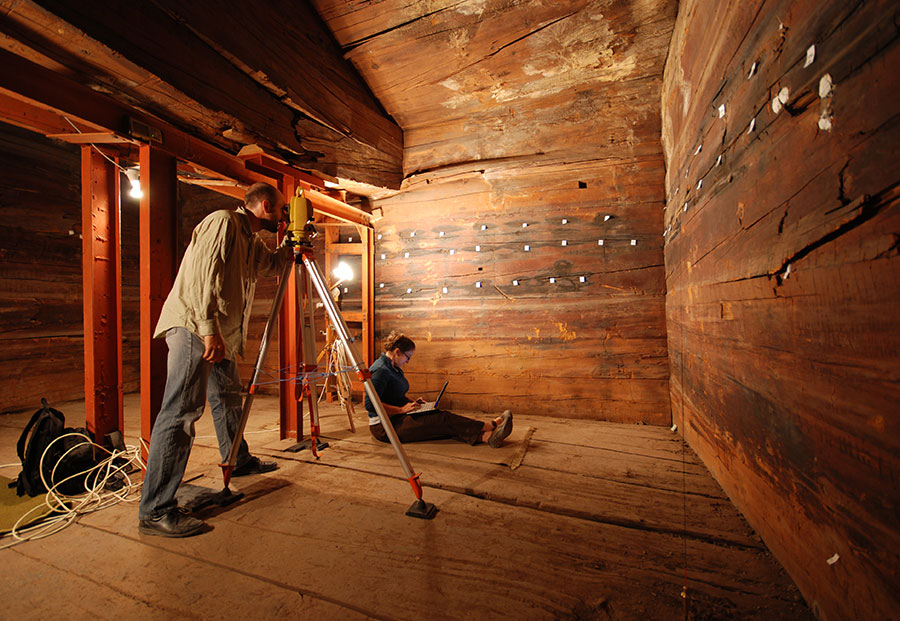 A person using survey equipment to record room features, another person sits on the floor.