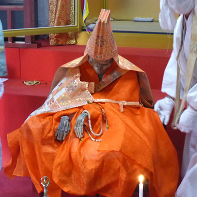 Mummified remains in a seated position, dressed in a bright orange robe and headpiece.