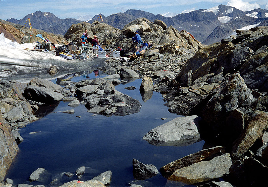A view of the excavation and team on a rocky and snowy mountain top, mountain peaks surrounding them in the distance.