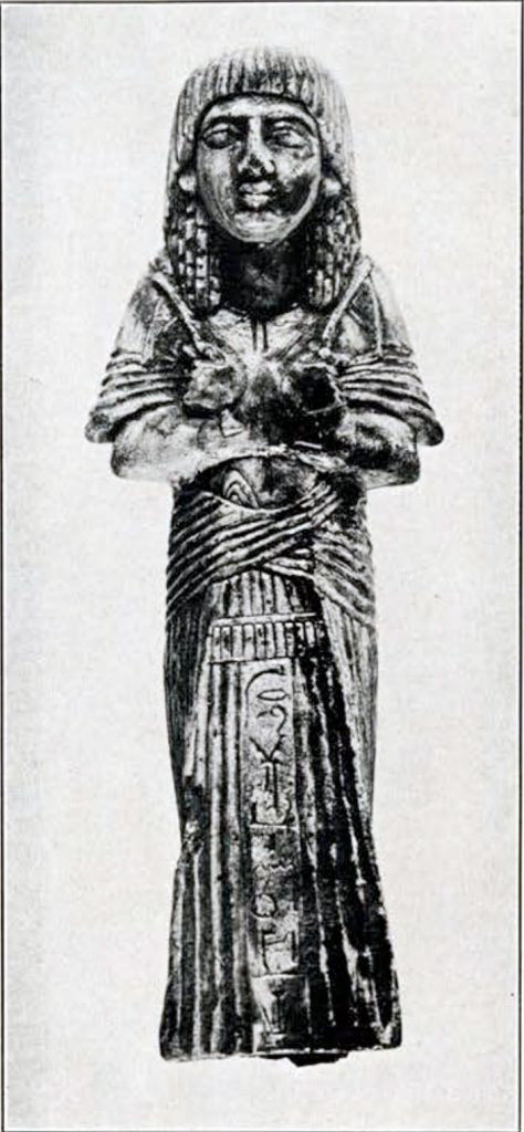 Black and white photo of statuette, a person with arms crossed and held close to the chest wearing a long gown. Hieroglyph inscription down front.