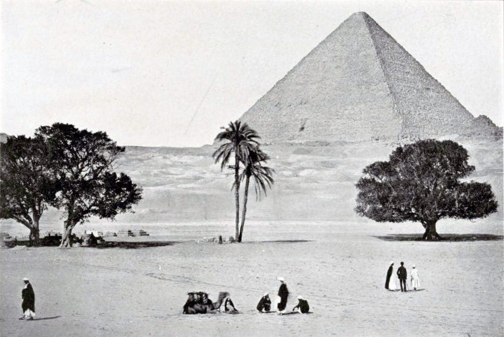 A pyramid at Giza with the desert surrounding it, three trees and some people wandering about, one with a camel