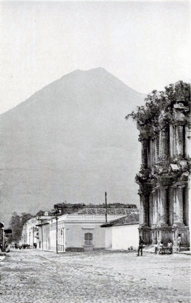 Part of a overgrown palatial building with two stories of columns and other, newer buildings lining a street, with the volcano De Agua rising in the distance