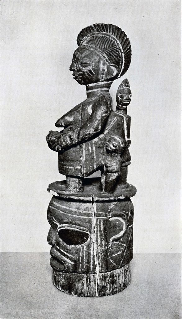 A carved wooden mask of a woman with other small figures, standing atop a head, in profile