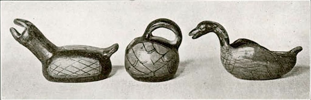 Three different small objects, two on either side figures that resemble birds and a small jar with a handle in the middle