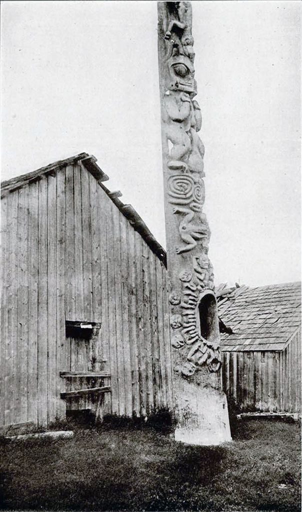 A totem pole in situ at Kit-wan-kool in front of buildings
