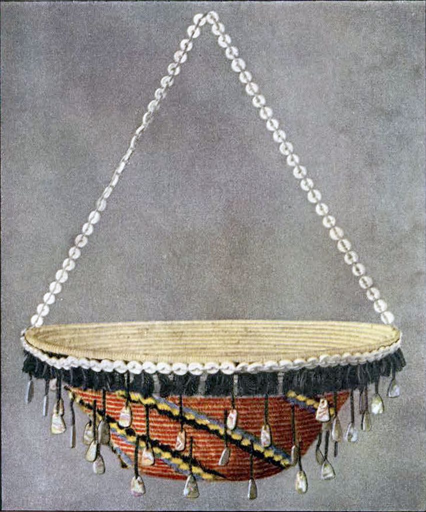 A suspended red basket with blue, black, and yellow stripes, a black feathered lip, and dangling shell beads
