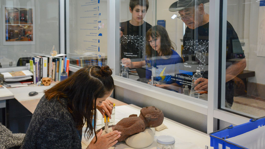 A conservator working on an artifact while a family looks on.