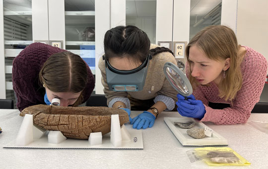 Three student curators in a lab, wearing latex gloves, and examining an object.