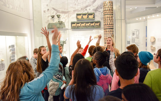 A school group in the Middle East Galleries, with hands raised to ask a guide questions.