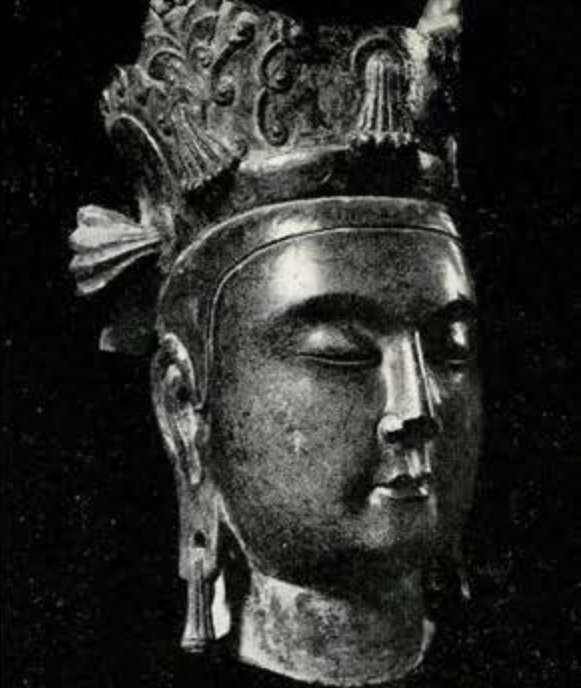 Stone head of bodhisattva, life size, narrow oval face with half-closed eyes and tiny mouth; no urna; long ears with pierced lobes and earrings; three peaked headdress with tassels partly missing
