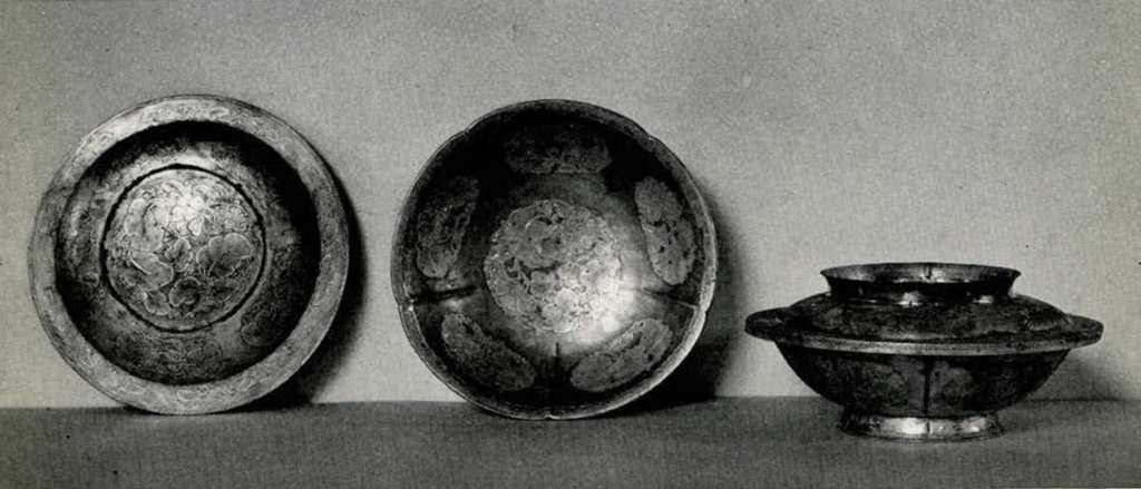 Two metal bowls, with lids, one standing with lid on, the other tilted to with the lid off to show the interior