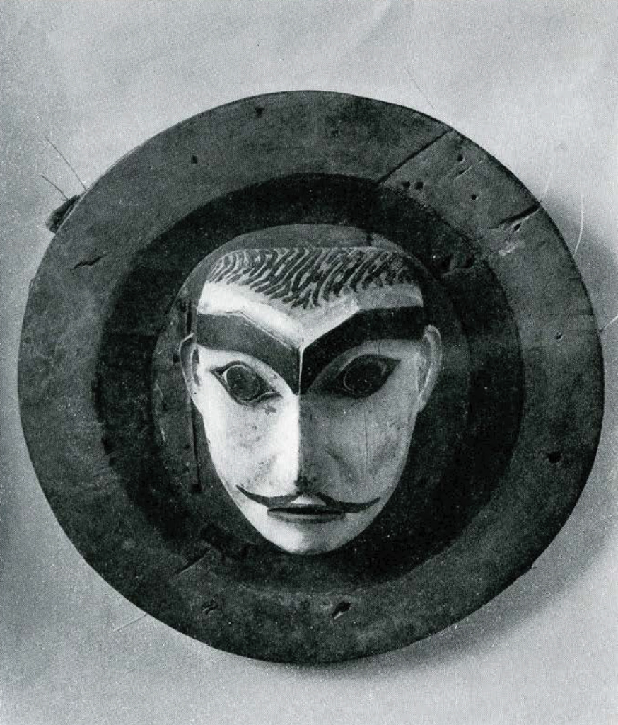 Circular mask with protruding painted face with thick unibrow and thin moustache.