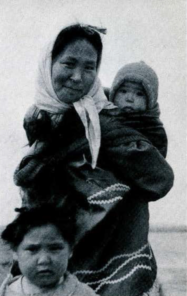 A mother holding a bundled child, another child stands in front of her.