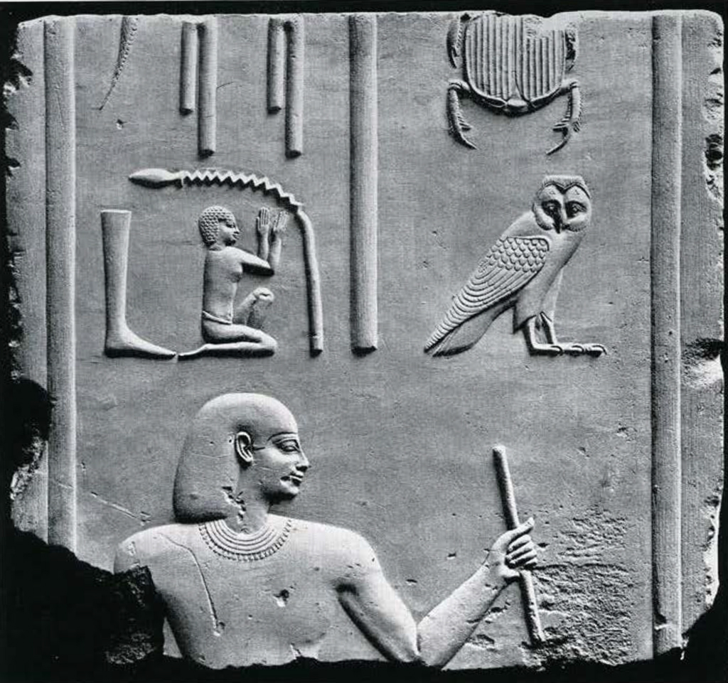 Relief of a man facing right wearing a bag wig and collar, he holds a staff, above him are two fragmentary columns of very detailed hieroglyphs.
