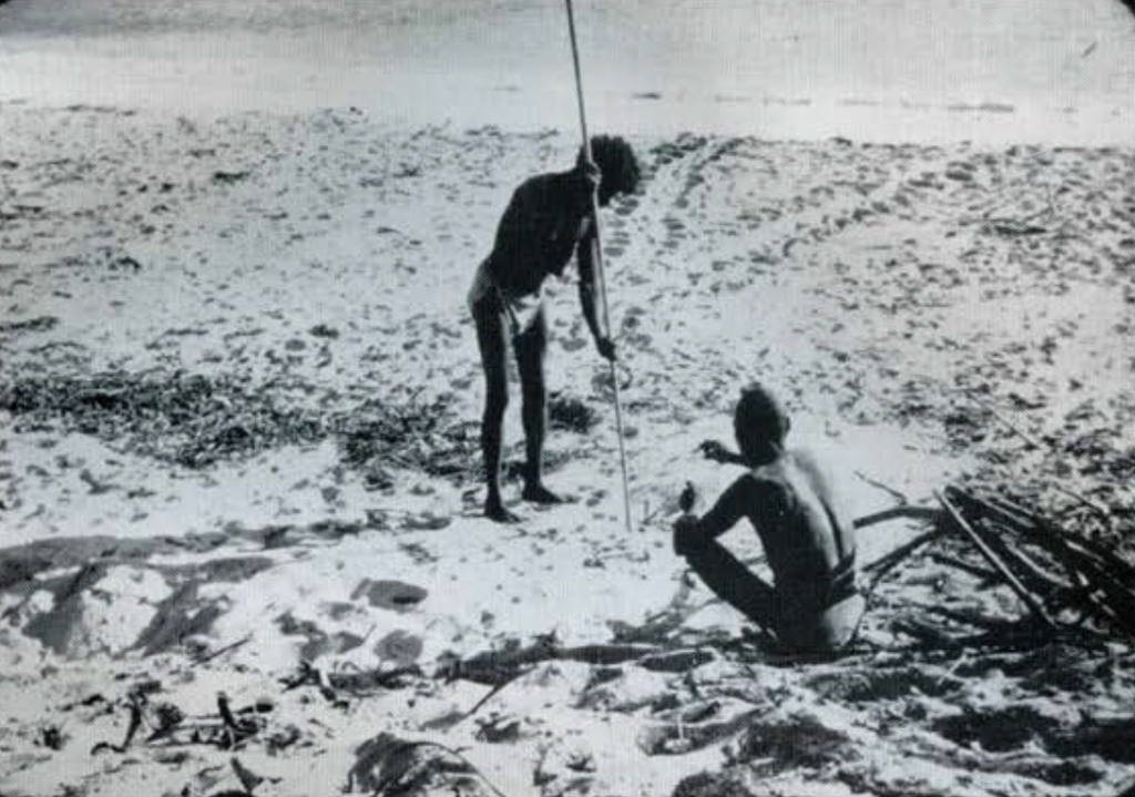 A man seated on the beach sand, another man with a long pole probing into the sand.