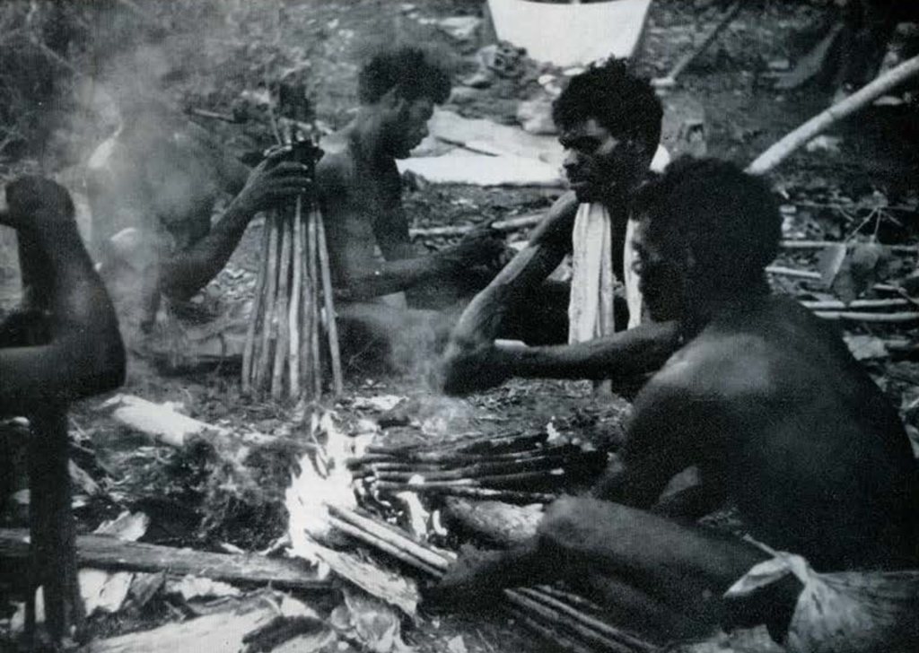 A group of men seated around a fire, holding budles of sticks near it.