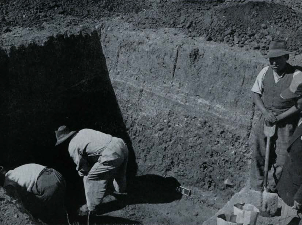 Two men digging in a rectangular pit while two other men watch, holding shovels.