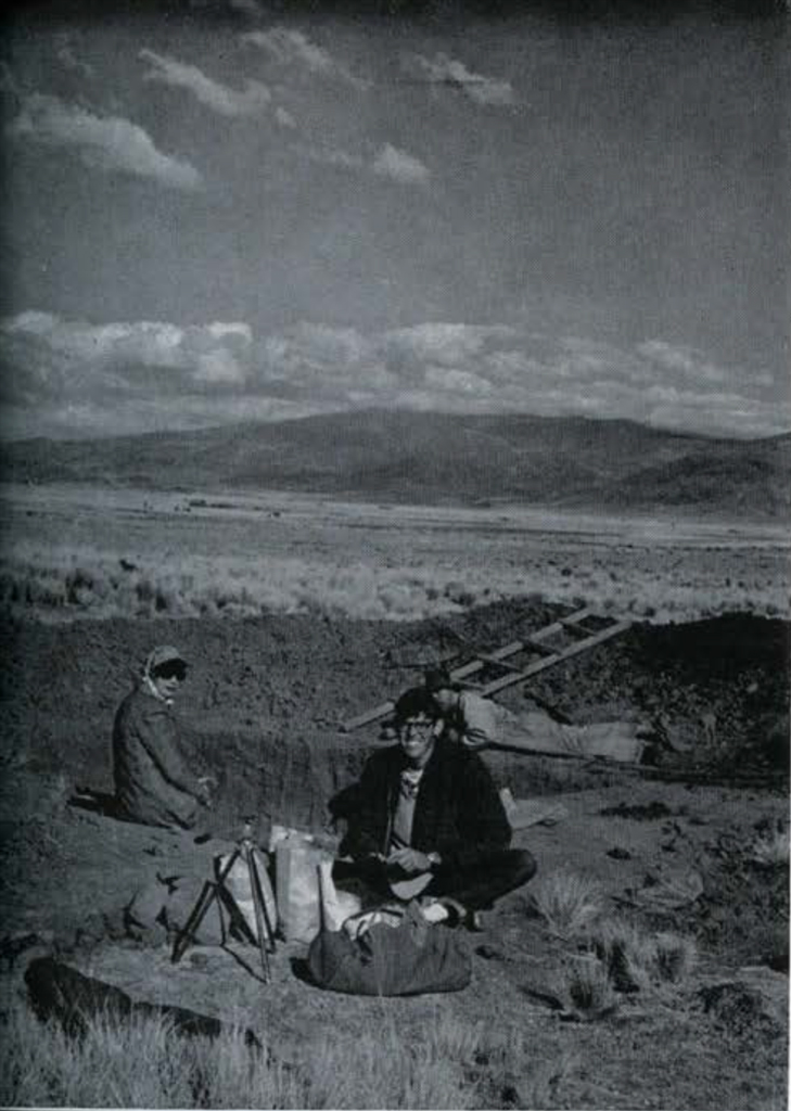 Two men sitting on the ground, the plains stretching into the distance behind them.