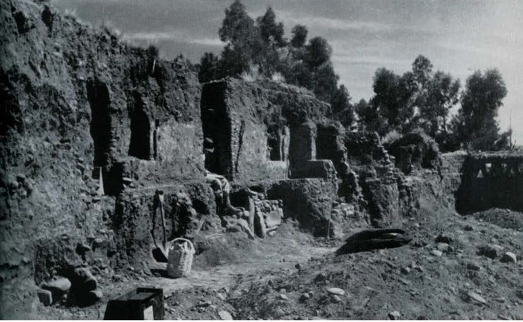 An excavated house with stone walls, windows, and a doorway which a man is sitting in.