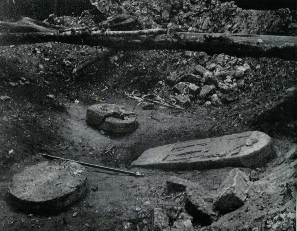 A stone stela and two round altars laying on the ground, a pit.