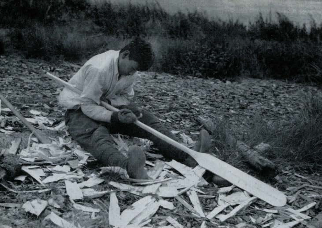 A man sitting on the shore surrounded by wood flakes, whittling a large piece of wood into a paddle.
