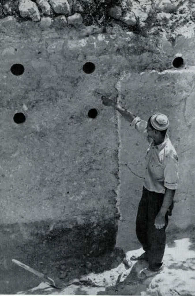 A wall with pottery embedded in it, a man pointing to holes in the wall.