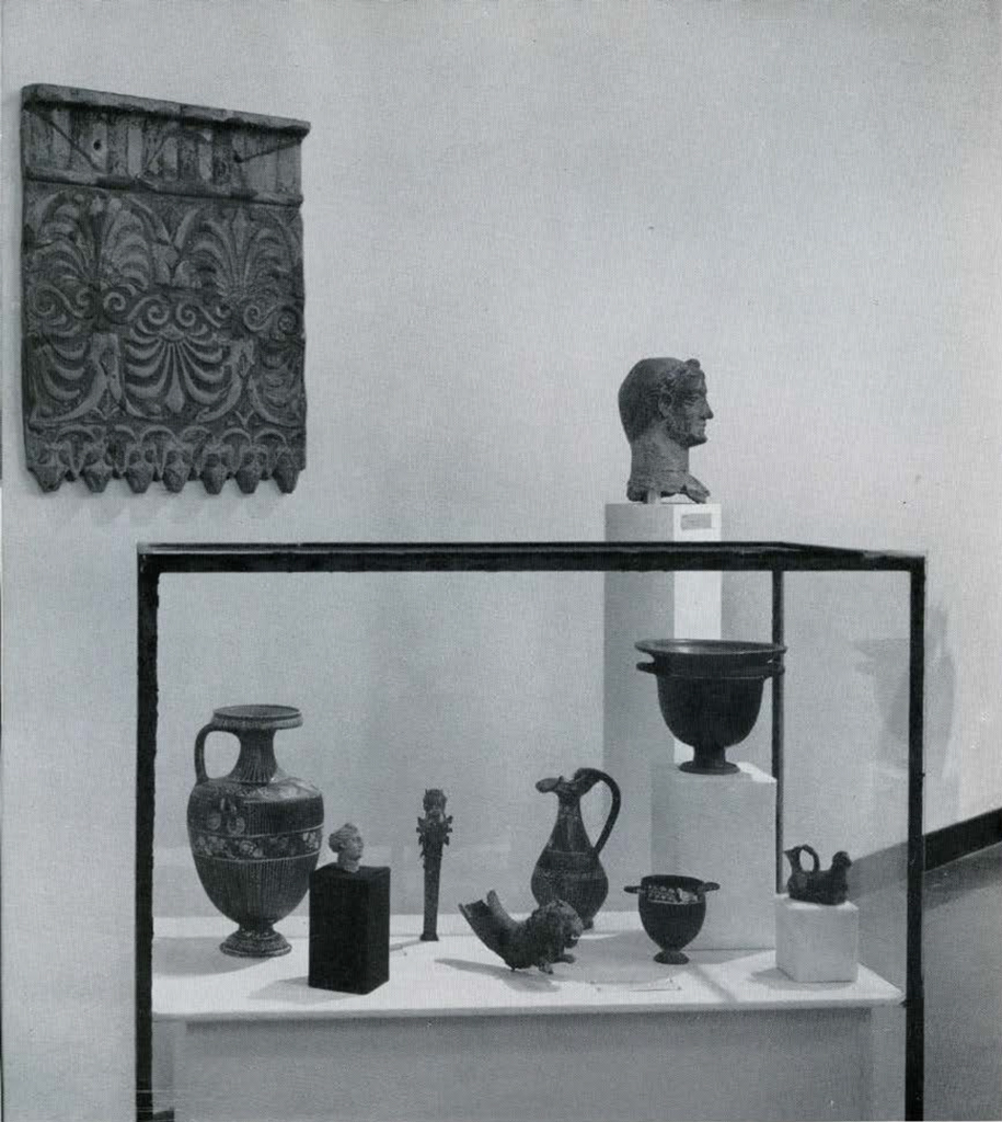 A variety of objects in a glass case, a bust on a pedestal behind.