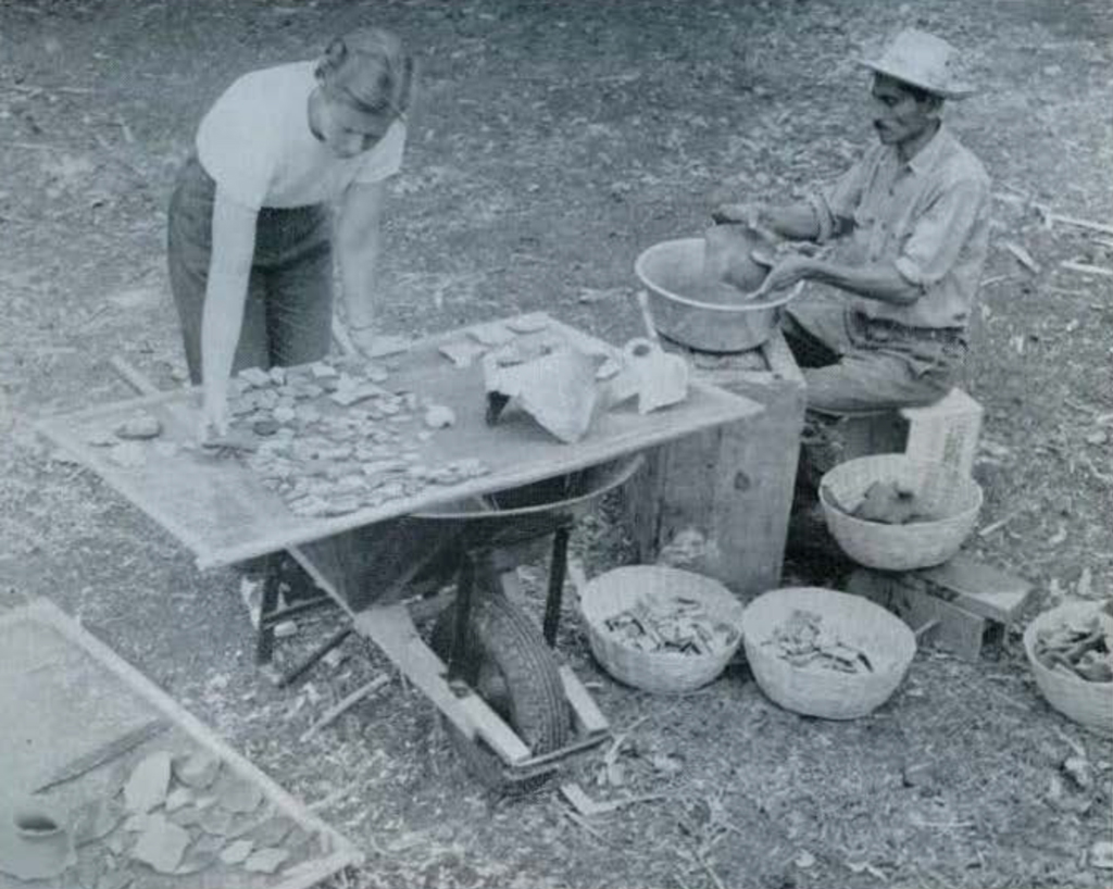 A man seated at a table washing pottery fragments in a basin, a woman sorting the fragments.