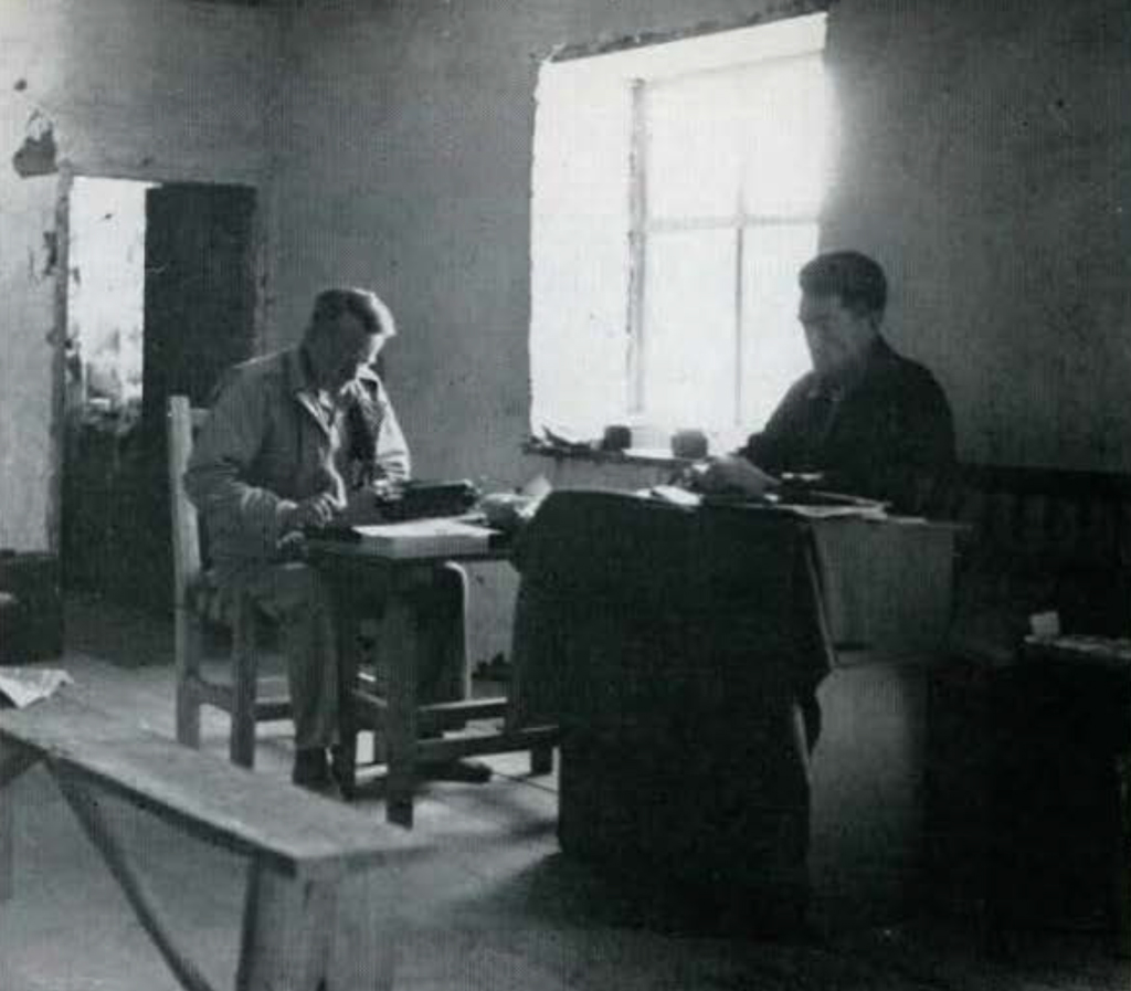 Two men seated at a table, reading.
