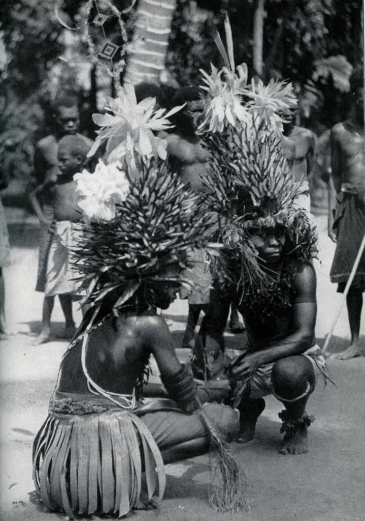 Two people squatting on the ground, wearing grass skirts and elaborate headdresses.