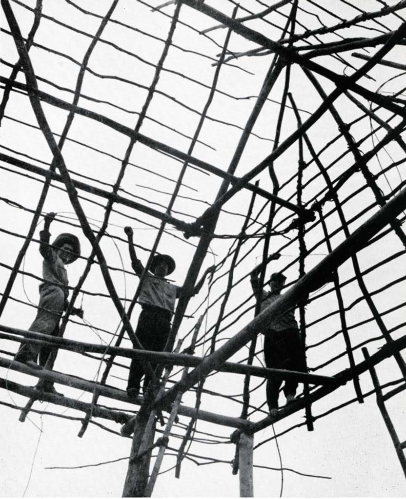 Three men standing on the tied together wooden frame of a roof of a house.